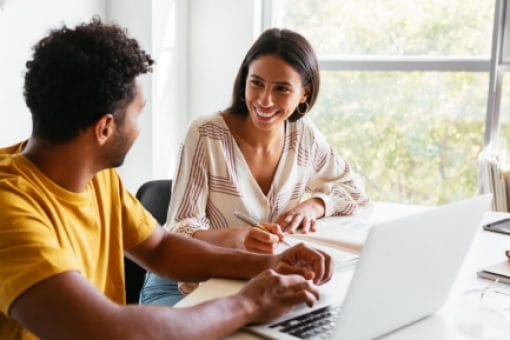 Two colleagues smiling and collaborating at a desk in a bright office, one working on a laptop while the other takes notes.