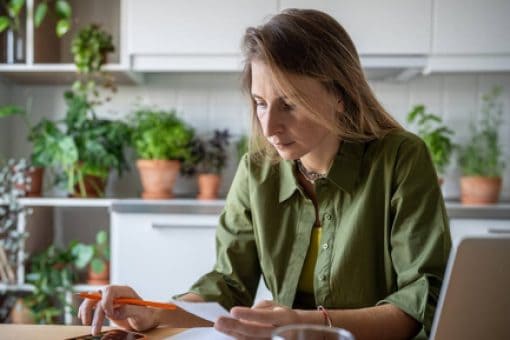 A woman uses a calculator on her mobile phone while holding a paper in her other hand. There's an open laptop on the table in front of her.