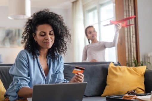 Woman uses a credit card while looking at her laptop as her teenage daughter plays with a toy airplane behind her.
