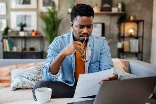 A man reviews a document in his hand with an open laptop in front of him on the table.