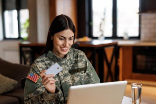 A woman in a military uniform sits at a table looking at a laptop while holding a credit card.