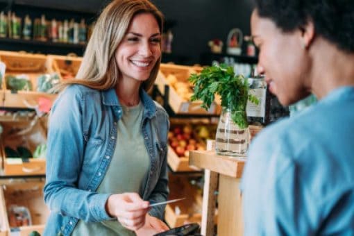 A woman in a produce market holds a credit card above a cashier's hand, who holds a payment terminal at checkout.