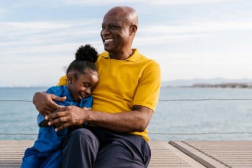 A man and young child laugh and hug as they sit together on a concrete bench overlooking water.