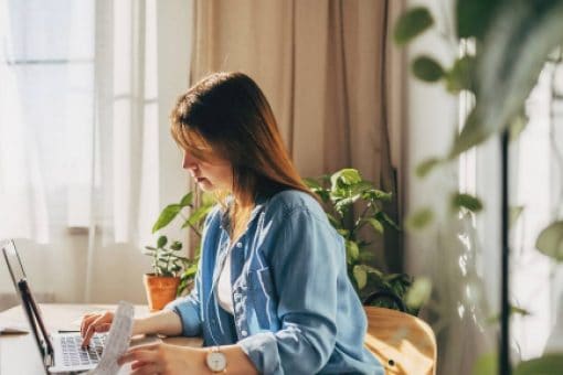 A woman sitting at a table types on her laptop with one hand and flips through paperwork with her other hand.