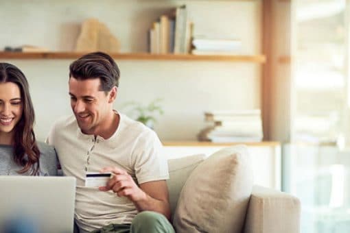 A couple sits together on a couch looking at a laptop. The man holds a credit card in his hand.