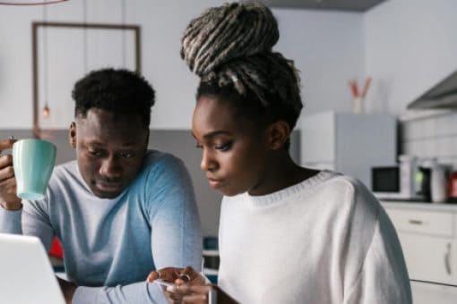 Couple sits at a kitchen table looking at a laptop. The woman points at the screen; the man holds a mug.