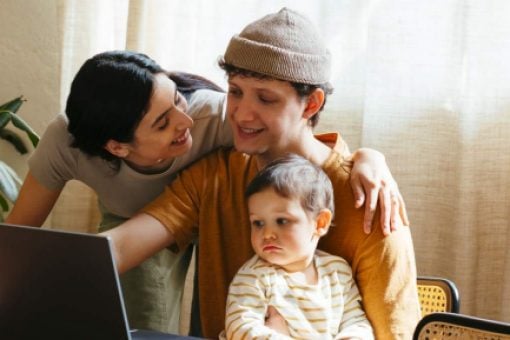 A man with a baby on his lap looks at a laptop as a smiling woman looks over his shoulder.