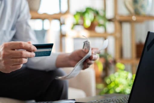 A person holds a credit card and some papers in their hand as they sit in front of a laptop.