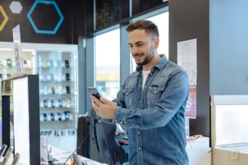 A man tests a smartphone for sale in a phone store, surrounded by displays of smart phones and other devices.