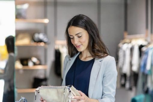 Smiling woman shopping in a boutique holding a brown handbag, surrounded by stylish clothing and accessories in a modern store setting.