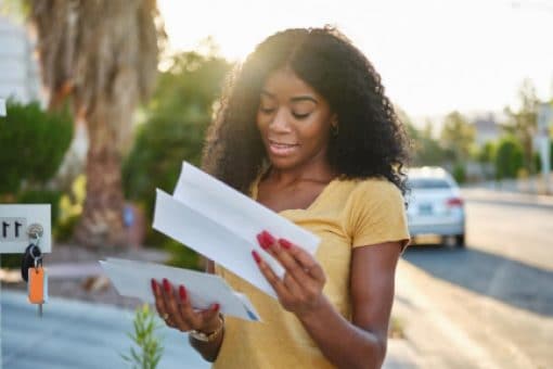 Woman checking her mail outdoors, smiling as she reads a letter near a community mailbox on a sunny day.