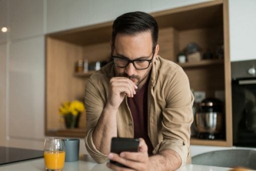 Man with glasses leans on a kitchen counter looking thoughtfully at his smartphone. A glass of orange juice is by his arm.