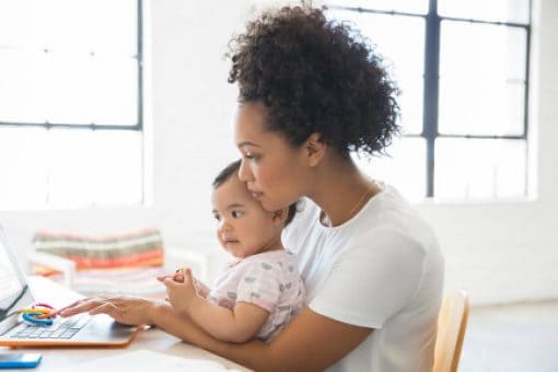 With a toddler in her lap, a young woman sits at a kitchen table and works on her laptop.