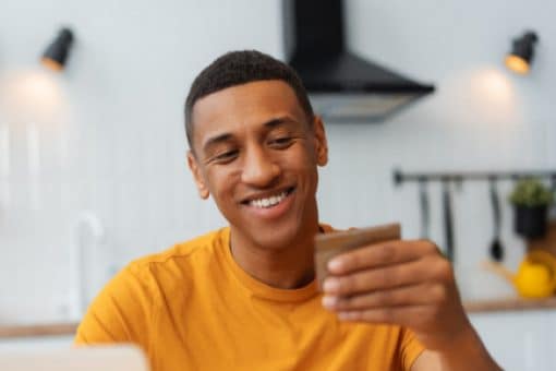 Smiling man in a yellow shirt holding a credit card while using a laptop in a modern kitchen