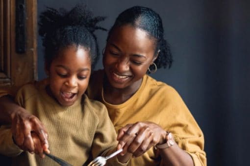 Smiling mother helping her young daughter cut food at a dining table, while another child sits beside them, all enjoying a meal together.