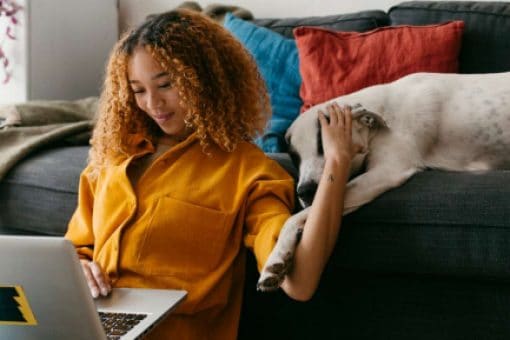A smiling woman sits on the floor in front of a couch, working on her laptop and petting a dog.
