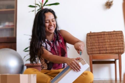 A woman sits on the floor smiling as she uses a tool to open boxes in her apartment.