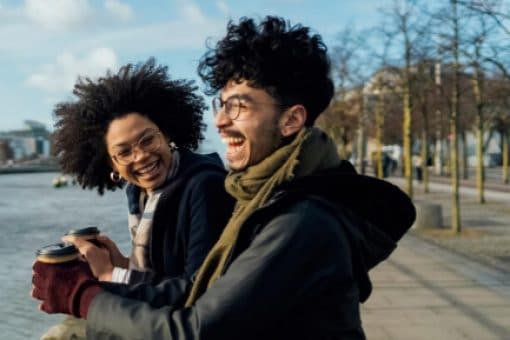 A man and a woman holding coffee to-go cups laugh while looking out over a river.