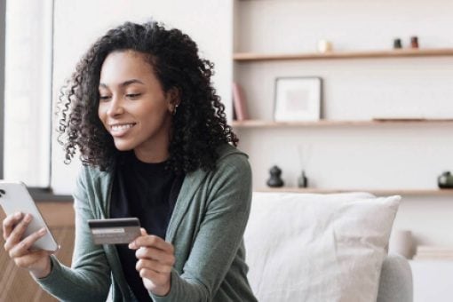 A woman sits on a chair looking at her mobile phone and holding a credit card in her other hand.