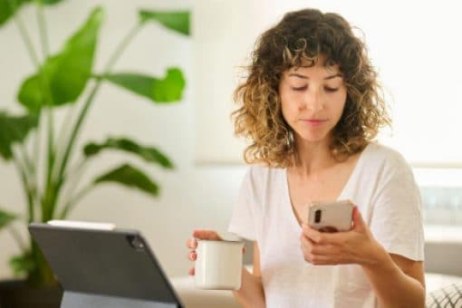 A woman sits on a couch holding a coffee mug and looking at her mobile phone.
