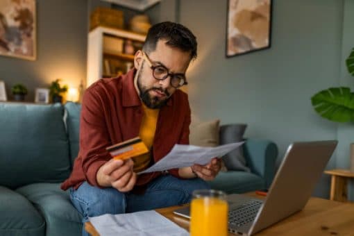 A man sits on a couch holding a credit card and his statement.