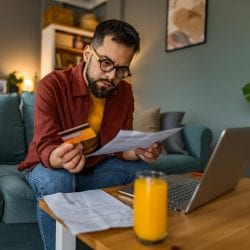 A man sits on a couch holding a credit card and his statement.