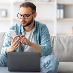 A man sits on a couch, scrolling on his phone in front of an open laptop.