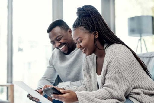 A man and woman sit together; he holds a piece of paper, she holds a mobile phone.