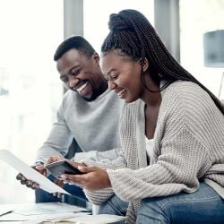 A man and woman sit together; he holds a piece of paper, she holds a mobile phone.
