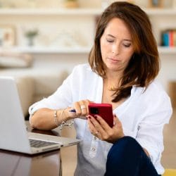 A woman scrolls on her phone and sits in front of a laptop reviewing her credit.