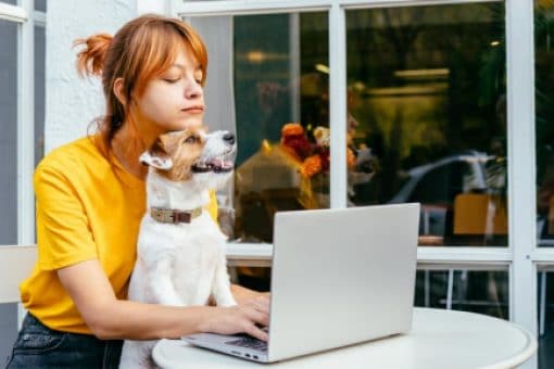 Woman sits outside with her dog in lap typing on her laptop