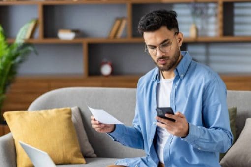 A man looks at his mobile phone in one hand while holding paper in his other hand.