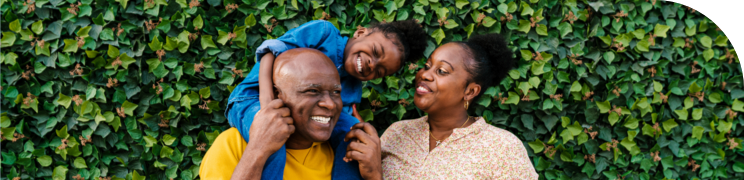 Smiling family with a young child on the father's shoulders, standing in front of a lush green ivy-covered wall