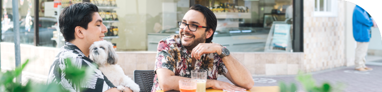 Two friends sitting at an outdoor café with drinks, one holding a small dog, while they chat and smile.