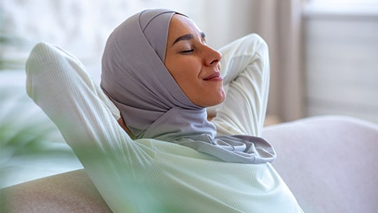 Woman in light blue hijab leans back on a couch with her hands clasped behind her neck and a content expression on her face.