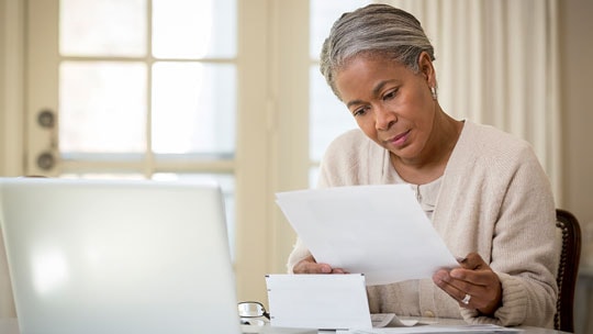 Woman sits at desk reviewing papers in front of a laptop.