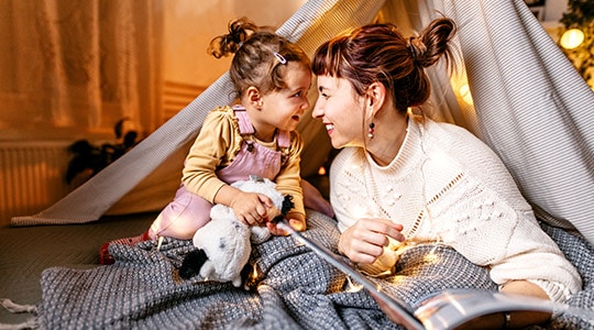 <Alt text> for image  max 6 words	A woman and small girl smile at each other while playing and reading.
