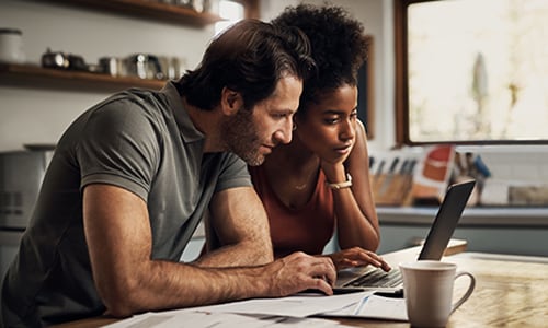 Man and woman sit at kitchen table looking at laptop.