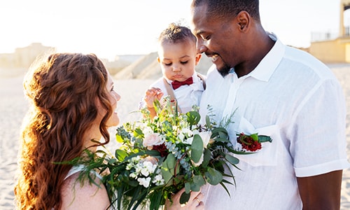 A smiling man and woman face each other as man holds a child and woman holds flowers.