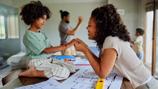 Woman leans on table covered with building plans and holds hand with little girl kneeling on table
