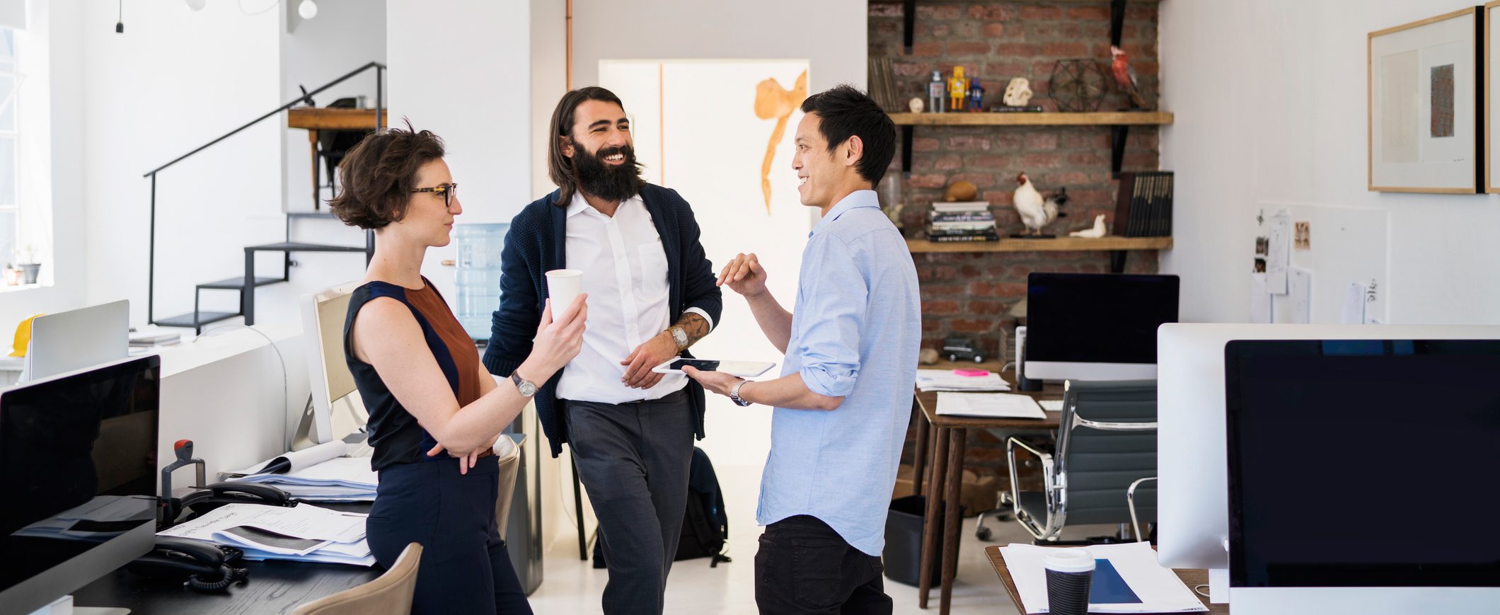 Three employees smile at each other in a workspace filled with desks and computer screens.