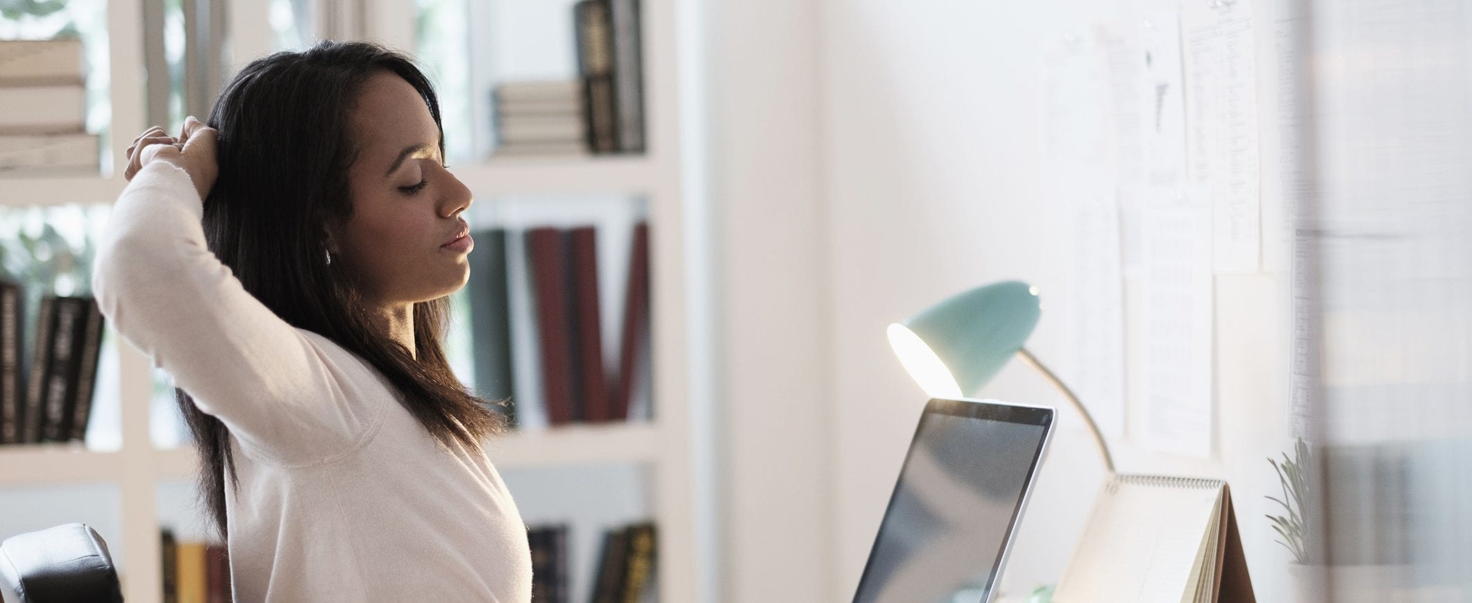A woman sits at a home office and stretches at her desk. 