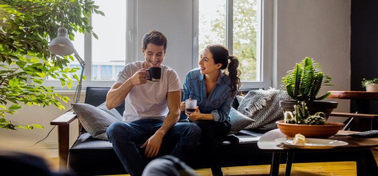 A man and a woman sit on a couch in their sunlit apartment. They are drinking coffee.