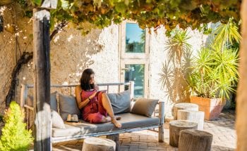 A woman wearing a red romper sits outside on a couch and looks at a tablet.