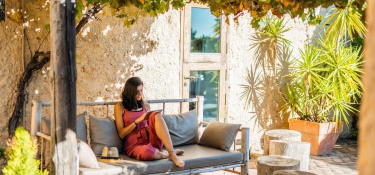 A woman wearing a red romper sits outside on a couch and looks at a tablet.