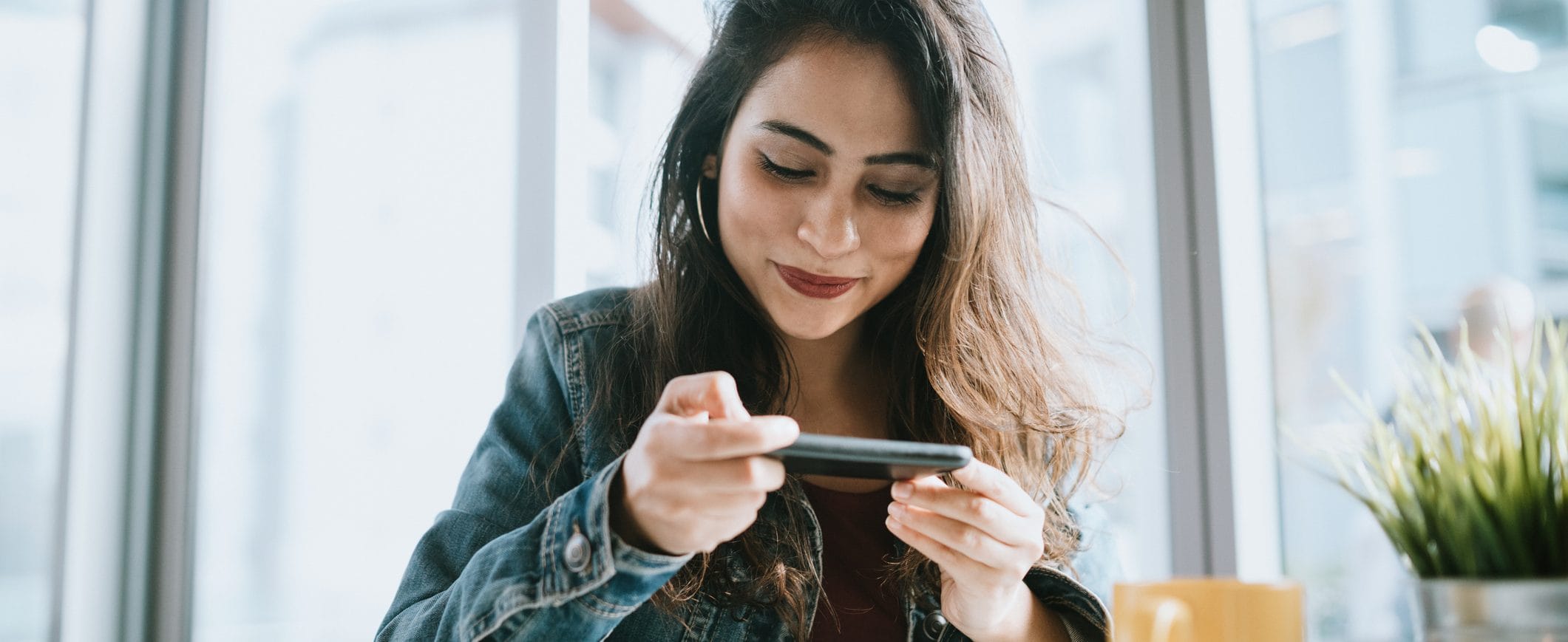A woman seated at a table with a yellow coffee mug, looking at her phone and smiling.