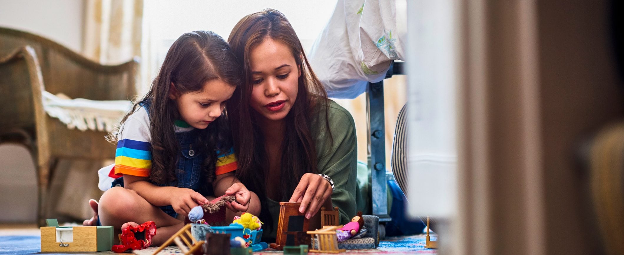 A mom and young daughter play on the floor with doll house furniture and figures.