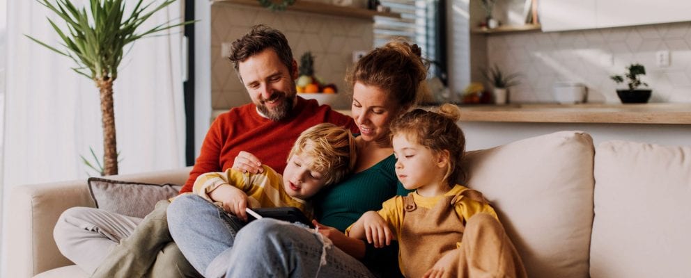 A family of four (mom, dad, a boy and a girl) sit on the couch and look at something.