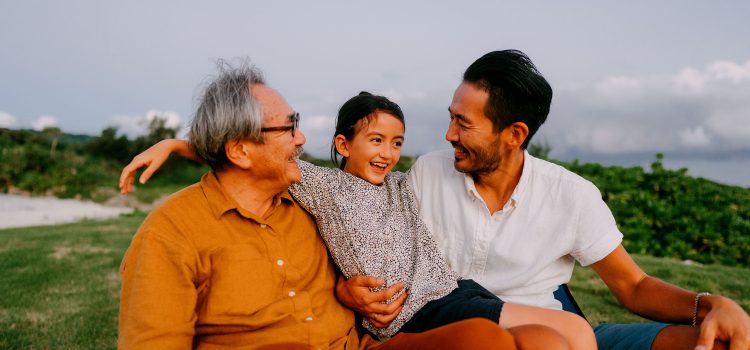 A grandpa, his son, and grandchild sit outside in the grass laughing.