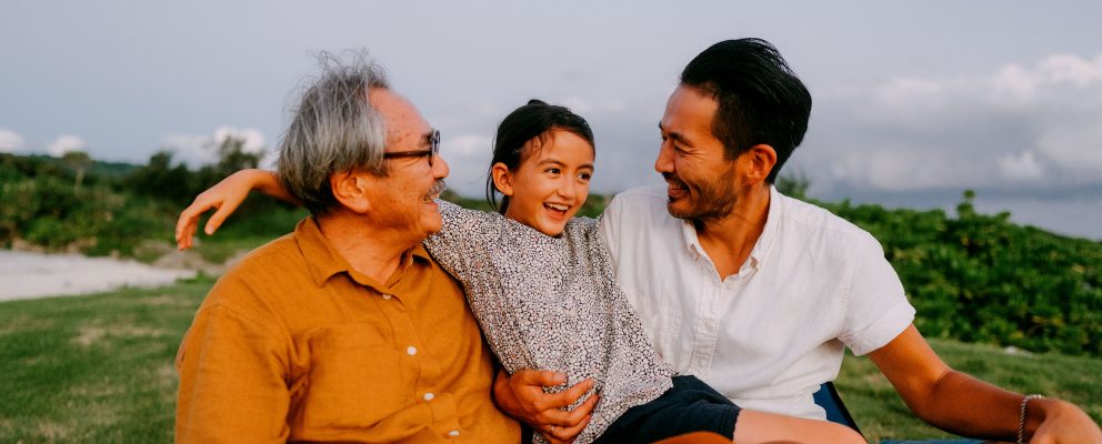A grandpa, his son, and grandchild sit outside in the grass laughing.